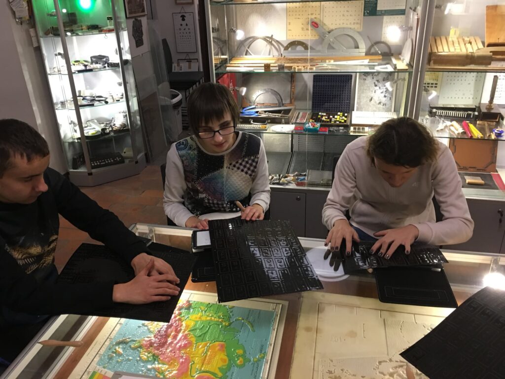 Kacper, Basia i Weronika przy podświetlonym stole ekspozycyjnym, na tle gablot w Muzeum Tyflologicznym podczas zapoznawania się z reliefowymi planszami przedstawiającymi kształty liter "widzących".