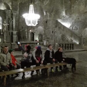 Grupa dzieci siedząca na długiej ławce w monumentalnej, podziemnej sali kopalni w Wieliczce. W tle ołtarz i ściany podziemnego kościoła oświetlone jaskrawo światłem z wielkich żyrandoli.