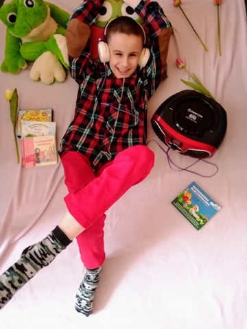 Kacper leży w kolorowych ubraniach na łóżku. Ma założone słuchawki a obok niego znajduje się odtwarzacz CD, z którego chłopiec słucha "Bajek z przesłaniem". Wokół Kacpra znajdują się sztuczne tulipany, bajki CD oraz maskotka żaby.