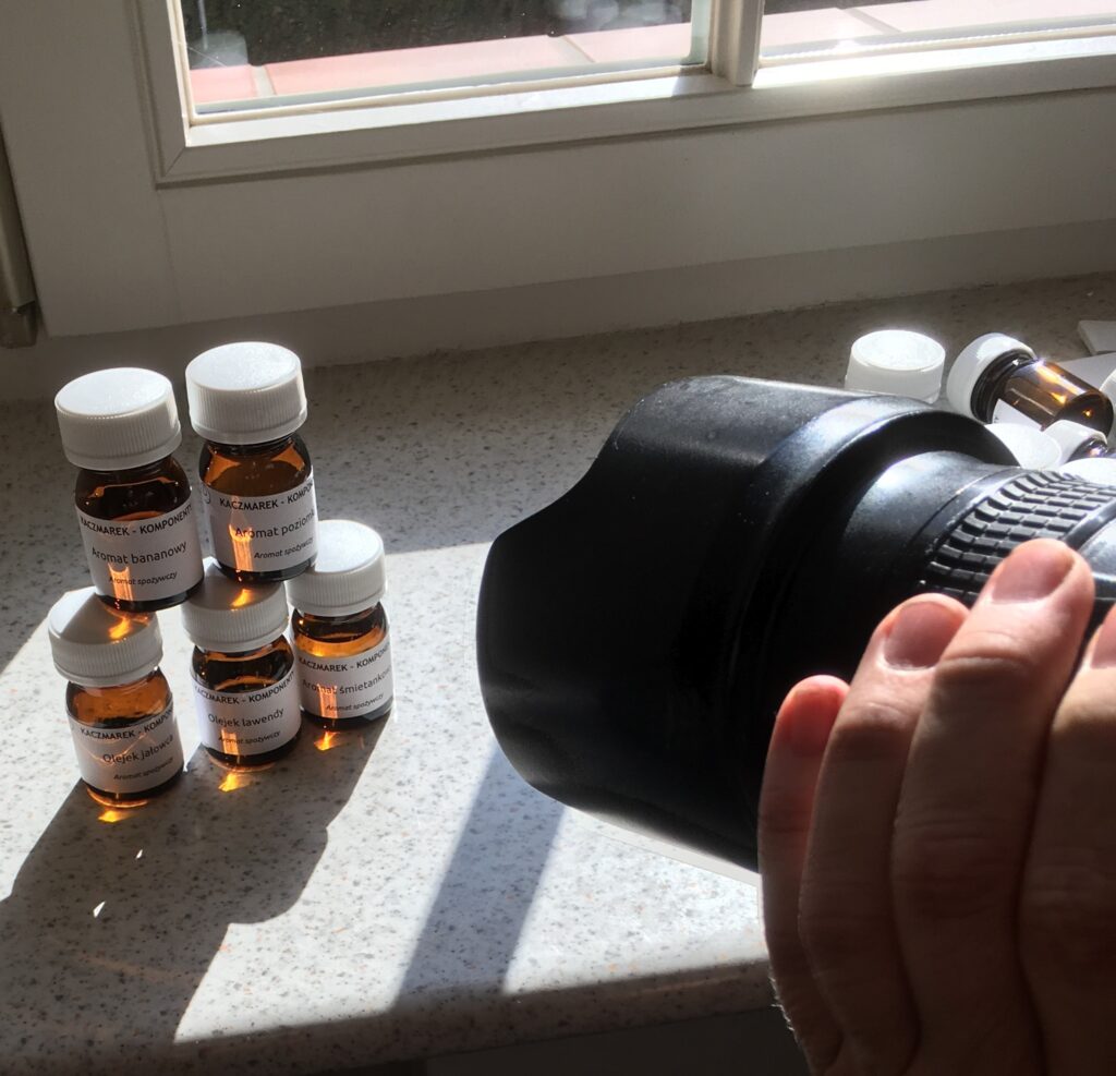 Obiektyw aparatu fotograficznego nakierowany na pięć buteleczek z aromatami stojącymi na parapecie okna do wirydarza.