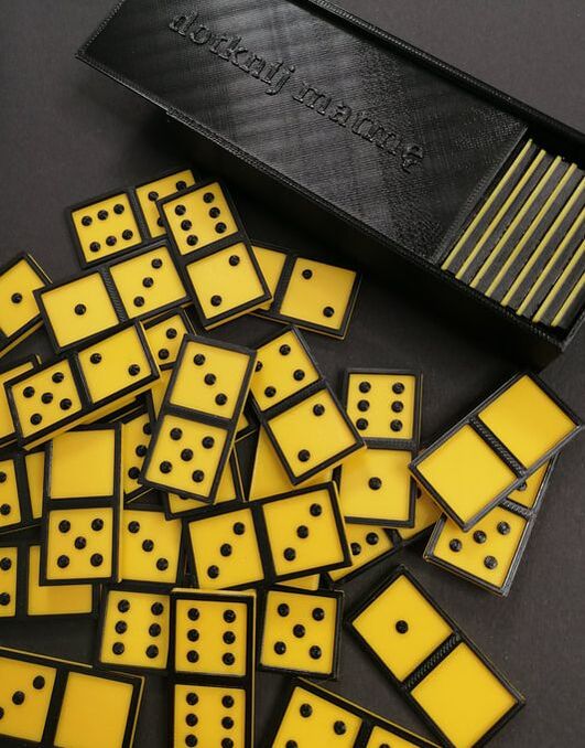 Domino żółte – rozrzucone kostki domina w kontrastowych kolorach: żółte tło, czarne obramowanie oraz wypukłe kropki. W prawym górnym rogu pudełko z wysuwanym wieczkiem do przechowywania kostek z napisem czarnodrukowym dotknij matmę.