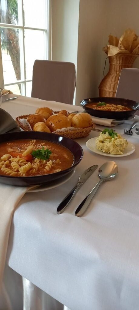 Na stole znajdują się głębokie talerze z nalaną do nich zupą pomidorową. Na środku stołu w koszyczku leżą pszenne bułeczki a na talerzyku znajduje się masełko czosnkowe.