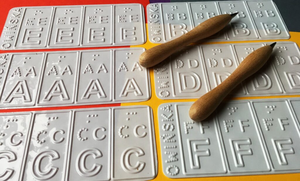 Tyflograficzne arkusze liter alfabetu łacińskiego do ćwiczenia pamięci mechanicznej wraz z dwoma kredkami do rysunku reliefowego. Inne ujęcie.