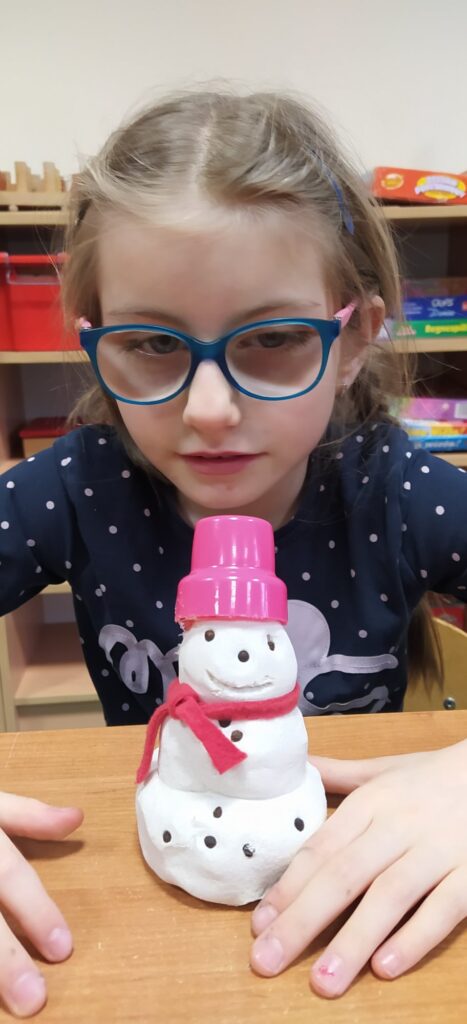 Dziewczynka siedzi przy szkolnej ławce, na której znajduje się wykonany z masy solnej bałwanek. Ma on na głowie różowy kapelusz z zakrętki od płynu do prania i czerwony szalik z materiału.