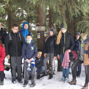Grupa zimowo ubranej młodzieży na zaśnieżonym szlaku wycieczki w Gorcach. W tle potężne pnie zielonych sosen.