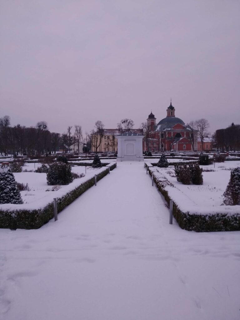 Widok na zaśnieżoną aleję ogrodu zmysłów. W tle biała kapliczka oraz budynek kościoła i Ośrodka.