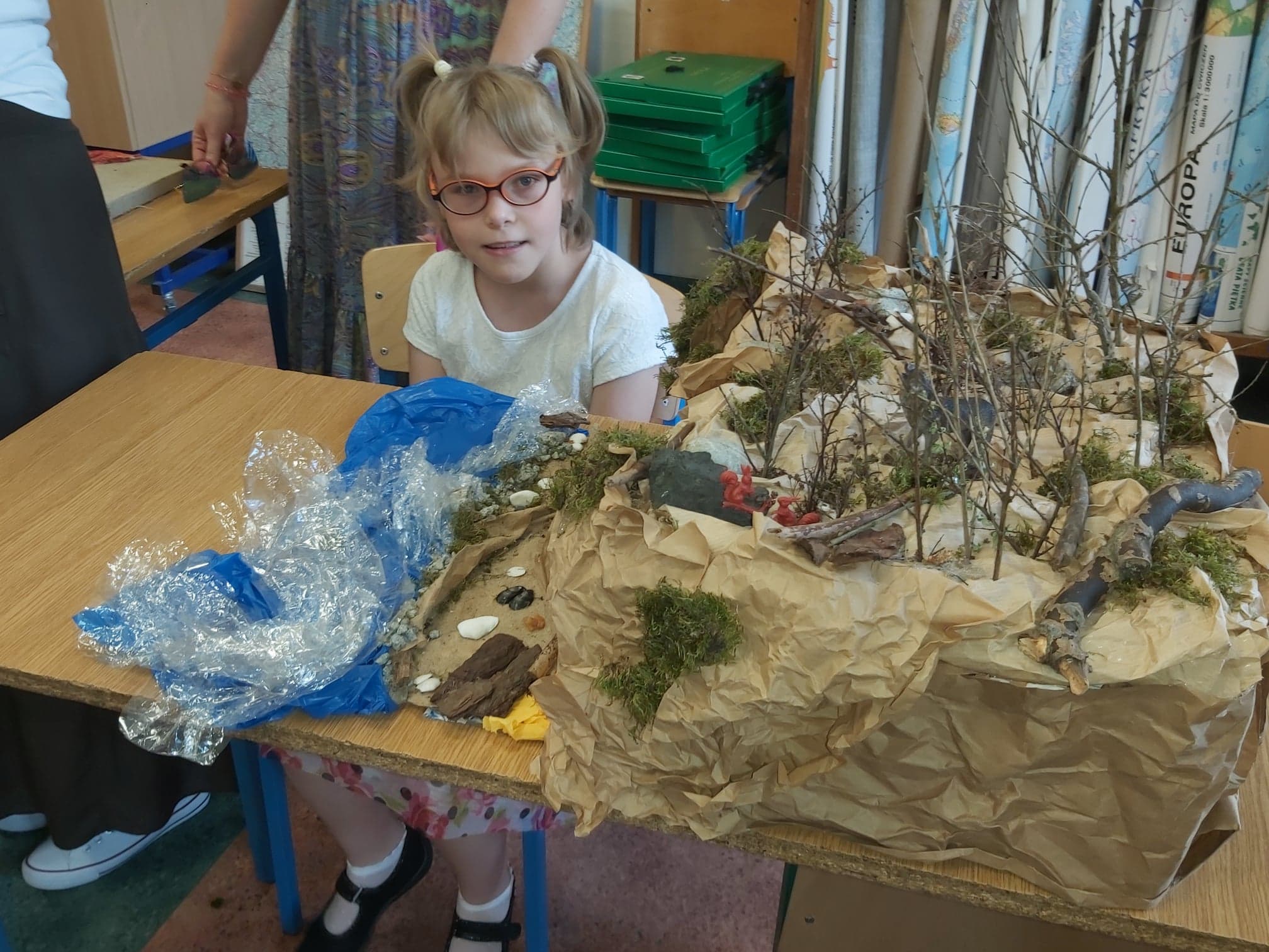 Kaja dumnie prezentuje swój model wybrzeża klifowego, w którym woda wykonana została z foli, na brzegu leżą kamienie, muszelki i kora, na klifie znajdują się drzewka wykonane z gałązek a nawet plastikowe zwierzęta (wiewiórki i wilk)