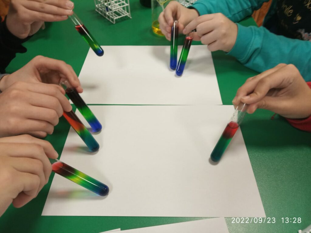 Na tle białych kartek uczniowie prezentują probówki z "tęczą".  W każdej probówce znajdują się kolejne warstwy cieczy różniące się barwą.