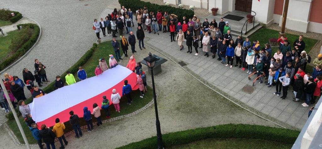 Grupa młodzieży trzymająca ogromną biało-czerwona flagę przed budynkiem Ośrodka. W tle zebrana młodzież i pedagodzy Ośrodka. Widok z lotu ptaka. 