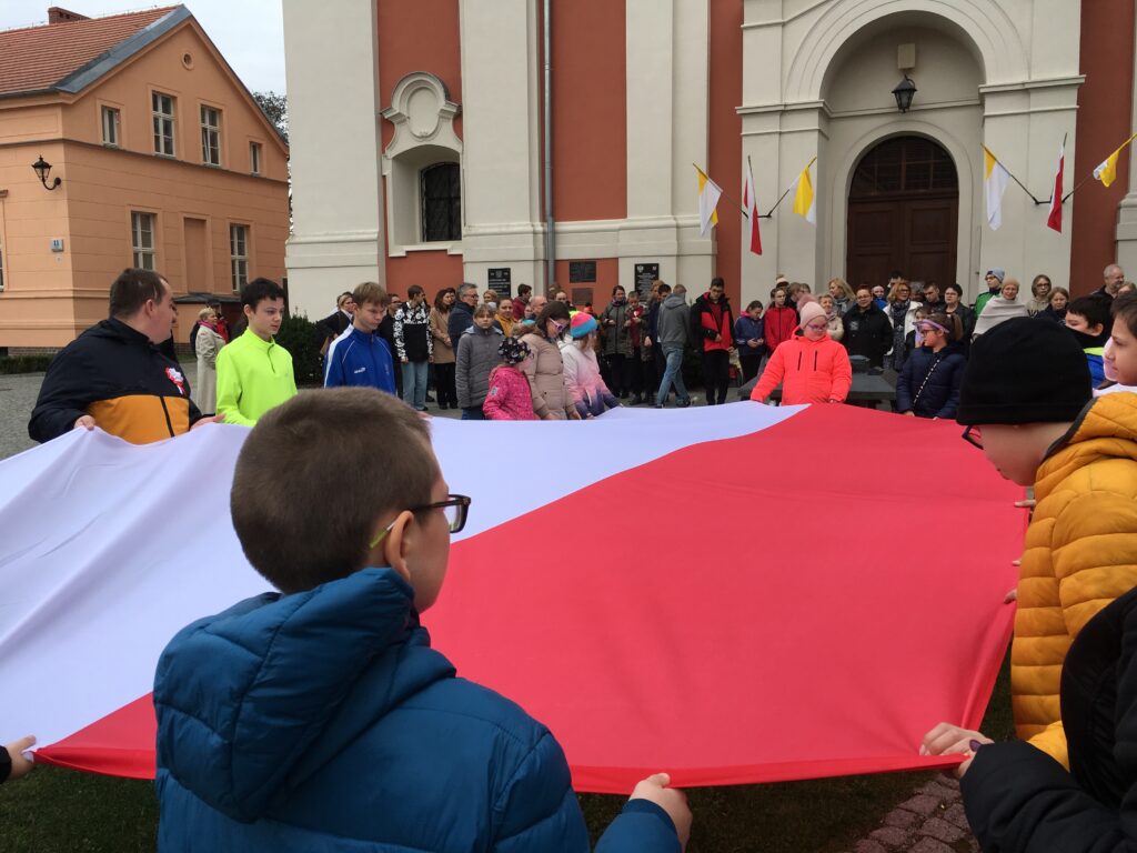 Grupa młodzieży trzymająca ogromną biało-czerwona flagę przed budynkiem Ośrodka. W tle zebrana młodzież i pedagodzy Ośrodka. 