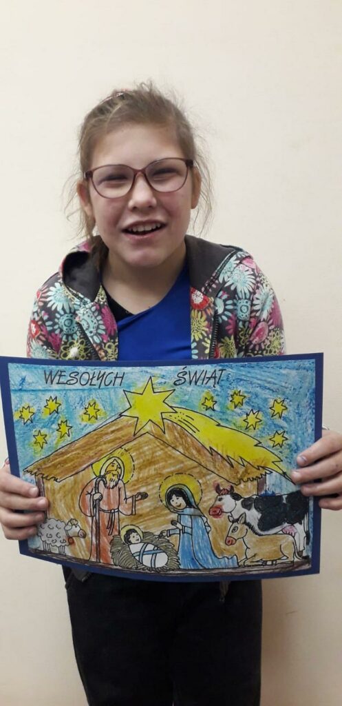 Ania prezentuje pokolorowaną przez siebie szopkę nad którą widnieją widnieją gwiazdy i napis ''Wesołych Świąt"