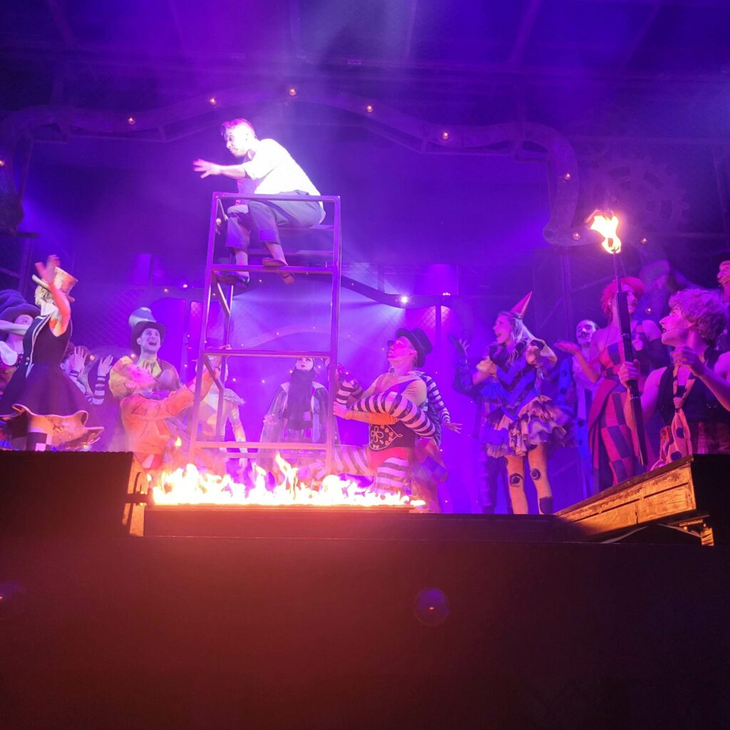 Jedna ze scen finałowych. Wszyscy aktorzy znajdują się na scenie. Główny bohater - Pippin siedzi na piętrowej, metalowej konstrukcji, pod nim płonie ogień.