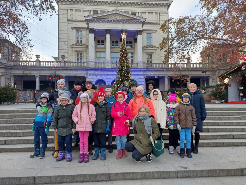 Grupa uczniów i nauczycielek na tle choinki i bożonarodzeniowych dekoracji znajdujących się na pobliskim Placu Wolności.
