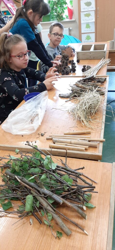 Dzieciaki przy biurkach. Na biurkach wcześniej posegregowane materiały: gałązki, bambus, kora, szyszki, siano.