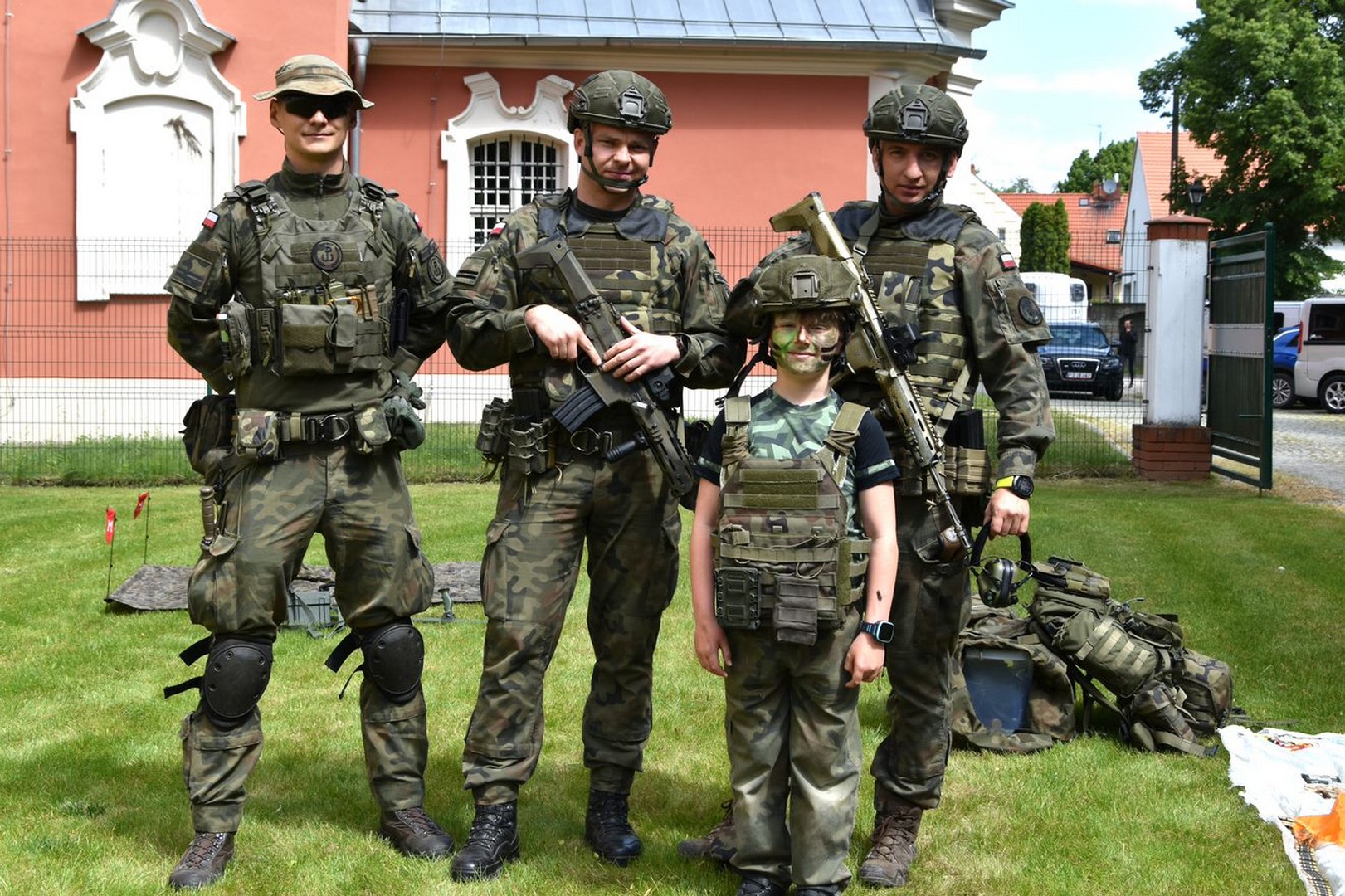 3 żołnierzy w mundurach i hełmach z karabinami stoją z chłopcem w przebraniu wojskowym.