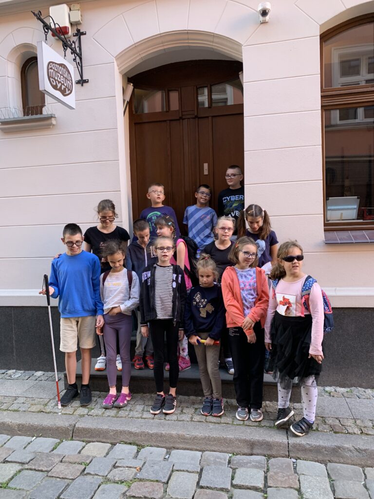 Grupa uczestników wycieczki stoi na schodach poznańskiego Muzeum Pyry. Od góry od lewej stoją: Jasiu, Kacper, Hubert, Jagoda, Kacper, Ewa, Kaja, Zuzia, Jasiu, Hania, Zuzia, Emilka, Marika i Liwia.