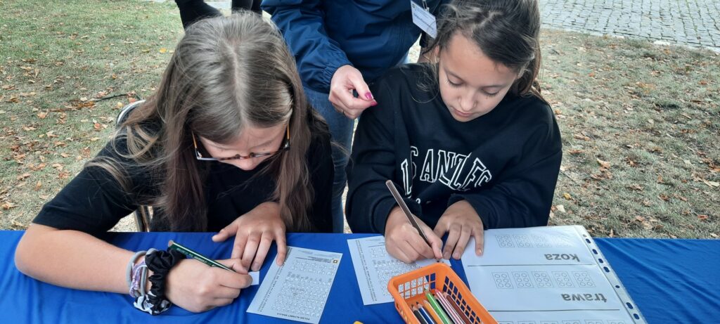 Dwie dziewczynki siedzą przy stole i odczytują przy pomocy alfabetu brajla zapisane przysłowia.