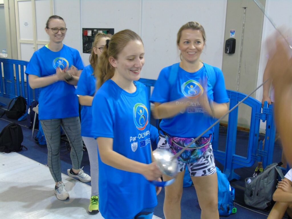 02. Uśmiechnięte Gosia, Marlena, Inga i pani Agnieszka stoją podczas pokazu szermierki. Wszystkie mają na sobie niebieskie koszulki z logiem imprezy paraolimpijskiej. Inga trzyma w prawej dłoni szpadę.