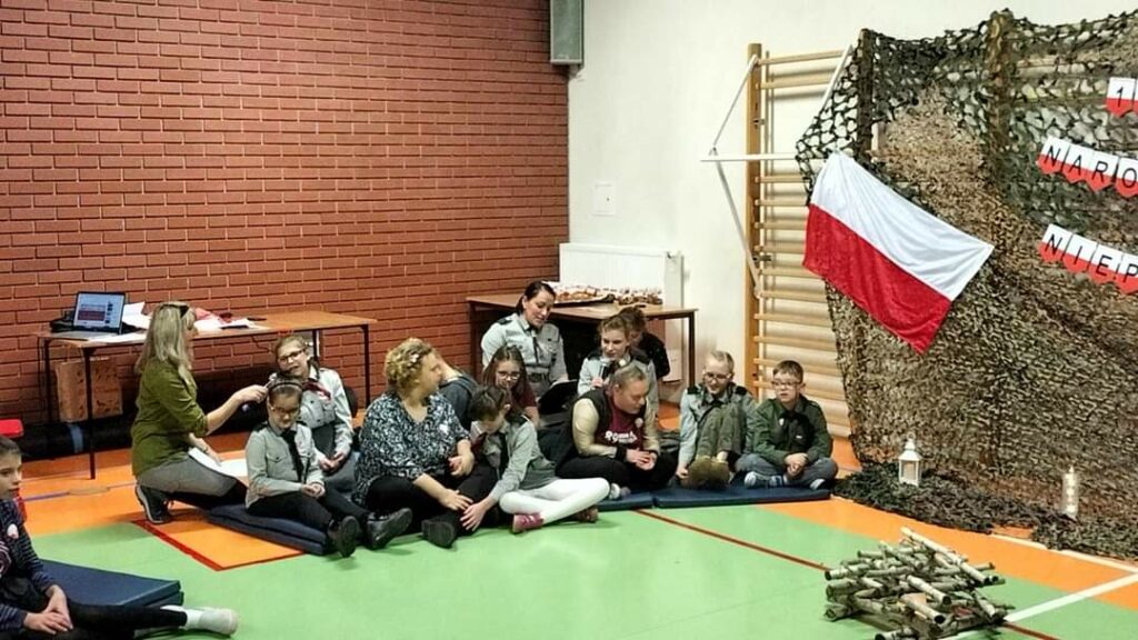 89 Wielopoziomowa Drużyna Harcerska Nieprzetartego Szlaku "Dreptacze" śpiewa piosenkę "Szara piechota". Harcerze siedzą na materacach przed imitacją ogniska.