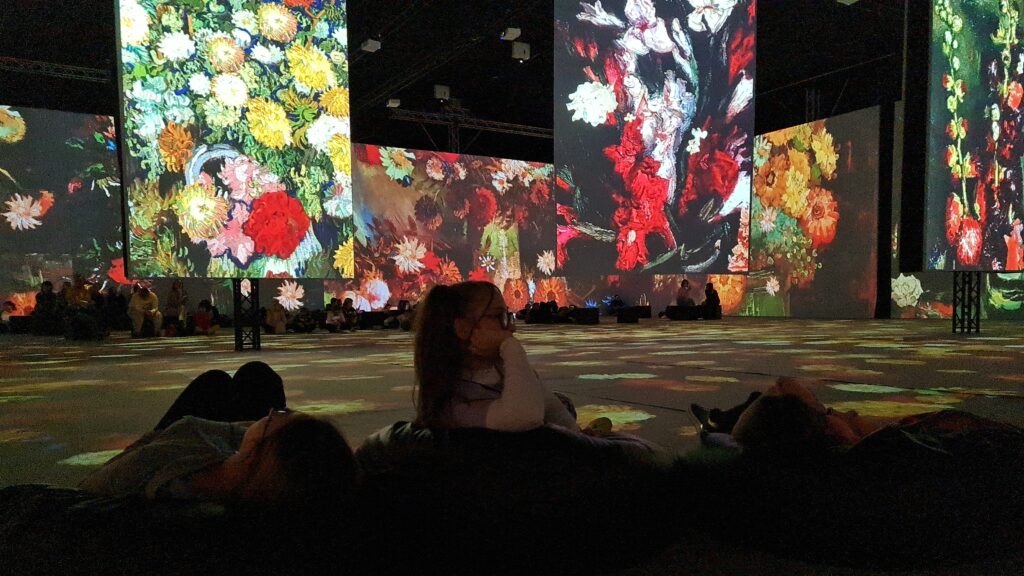 Dziewczęta leżą na pufach. Przed nimi na ścianach i na ekranach wyświetlane są obrazy Van Gogha przedstawiające kwiaty.