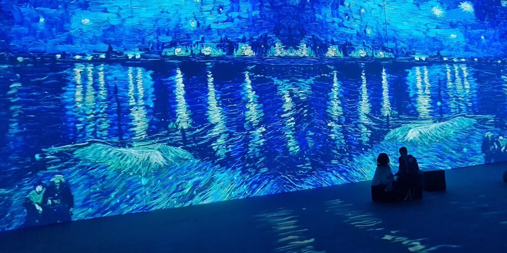 Na ścianie wyświetlony ogromny obraz "Gwiaździsta noc" autorstwa Van Gogha. Pani Dominika i Kacper siedzą na pufach obserwując grę świateł i obraz.