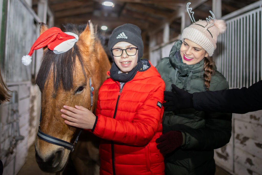 Zdjęcie zrobione wewnątrz stajni. Przy brązowym koniu Larrym stoi uśmiechnięty Janek, a za nim hipoterapeutka. Koń między uszami ma założoną czerwoną czapkę św. Mikołaja.