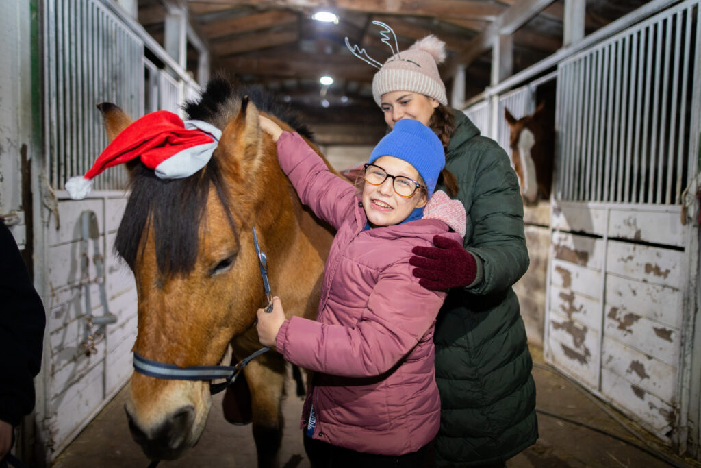 Zdjęcie zrobione wewnątrz stajni. Przy brązowym koniu Larrym stoi Ania, a obok niej hipoterapeutka. Koń między uszami ma założoną czerwoną czapkę św. Mikołaja.