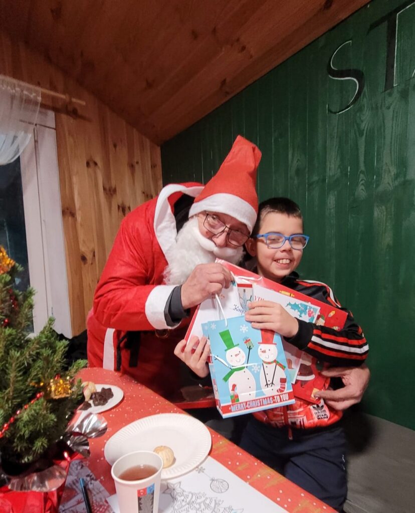 Zdjęcie zrobione w drewnianym domku. Do zdjęcia pozuje Kacperek z św. Mikołajem. Chłopiec trzyma w dłoniach prezenty.
