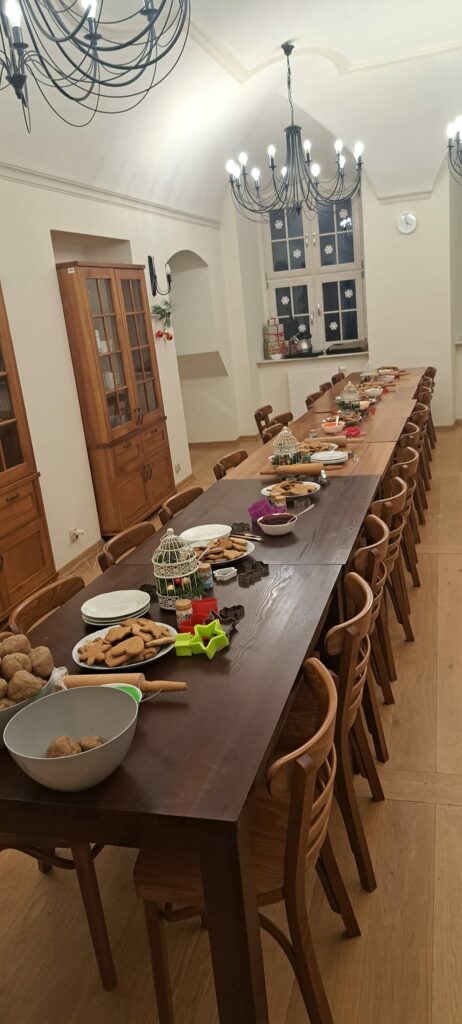 Mała stołówka. Drewniane stoły stoją złączone na środku stołówki. Na stołach leżą: miski z ciastem, talerze z upieczonymi piernikami, foremki do wykrawania pierników oraz miseczki z kolorowym lukrem.