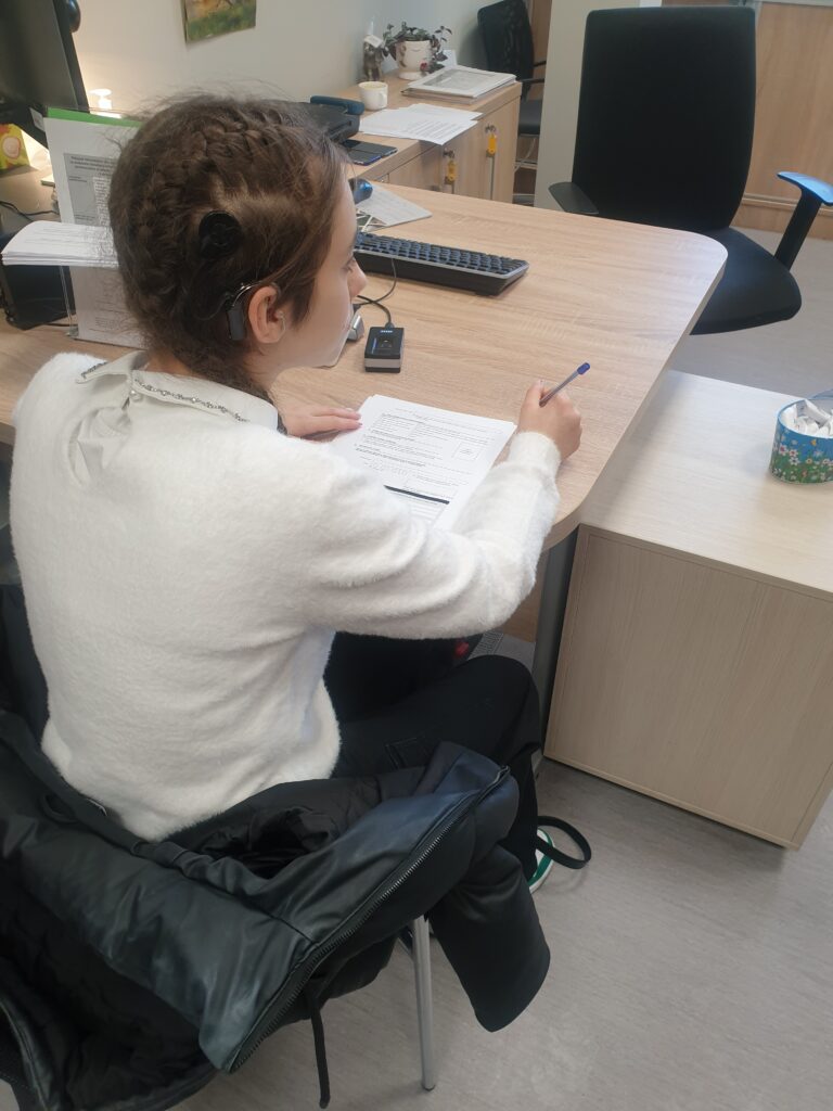 Dziewczyna siedzi przy biurku. W dłoni trzyma długopis i wypełnia leżący przed nią dokument.
