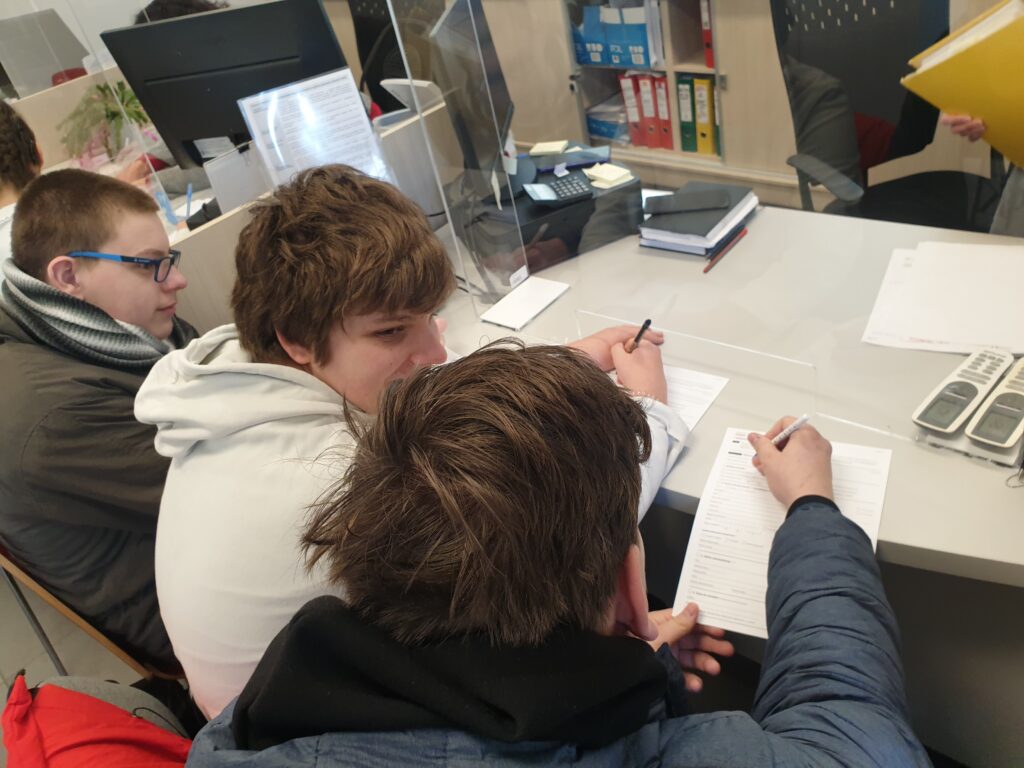 Trzech chłopaków siedzi przy biurku w urzędzie. Trzymają w dłoniach długopisy i wypełniają dokumenty.
