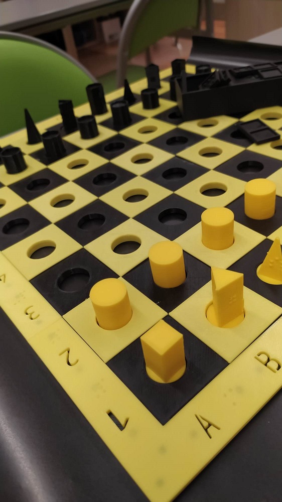Na stole rozłożona szachownica z ustawionymi pionkami do gry w szachy w kształcie brył. Szachownica oraz figury w kolorze żółto-czarnym.
