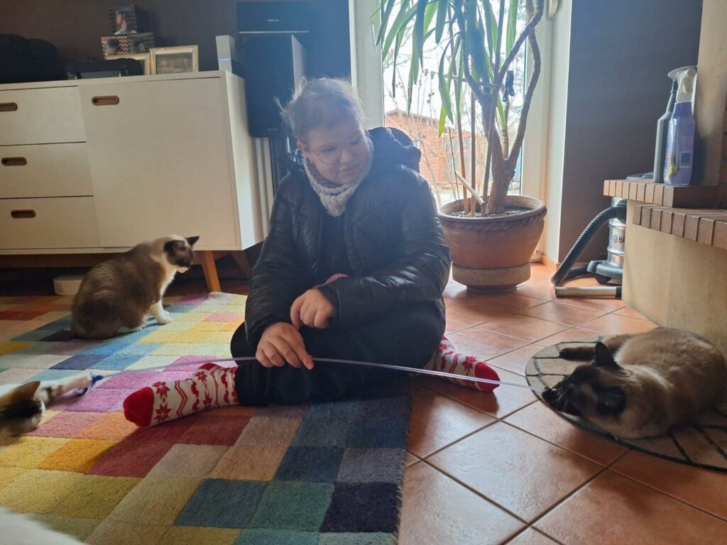 Nadia siedzi na dywanie. Obok niej trzy koty. W ręku trzyma zabawkę, którą zachęca kociaki do zabawy.