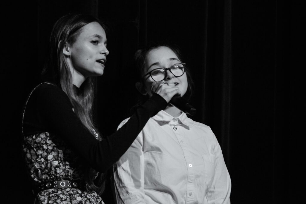 Czarno białe zdjęcie. Alina oraz Marlena wykonują wokalnie utwór. Alina trzyma w ręku mikrofon.