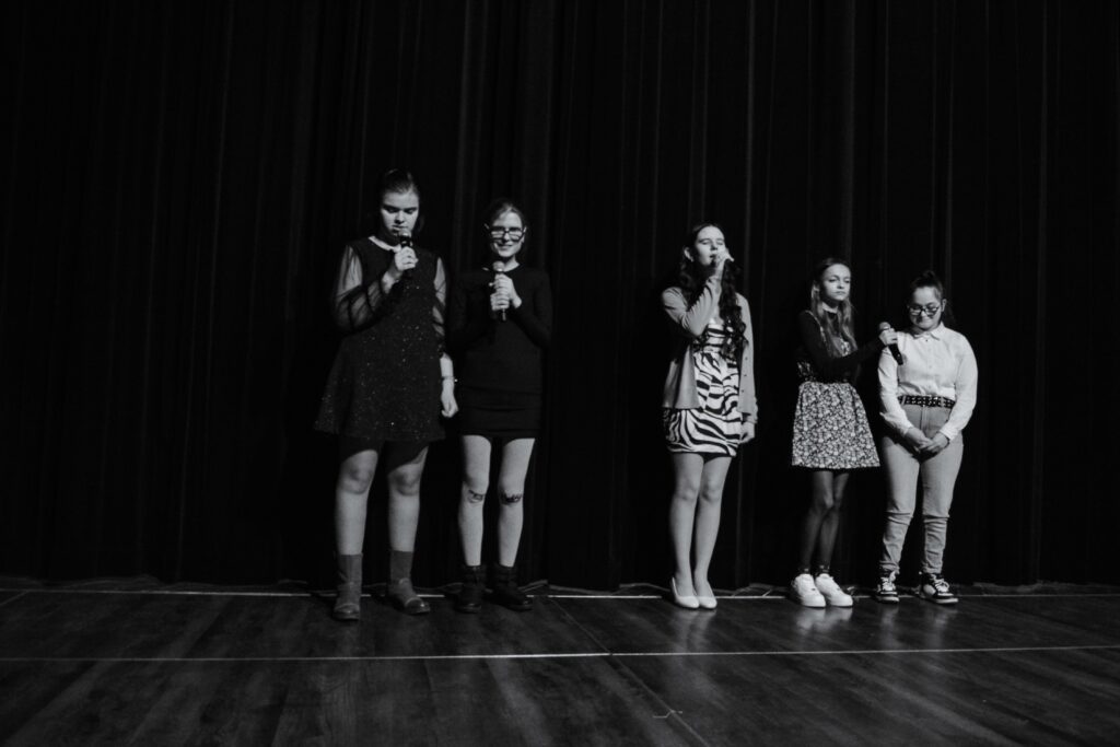 Czarno-białe zdjęcie. Uczennice stoją na scenie – od lewej: Zuzia, Jagoda, Kateryna, Alina, Marlena. Dziewczyny wykonują utwór. Z tyłu czarna, zasłonięta kurtyna.