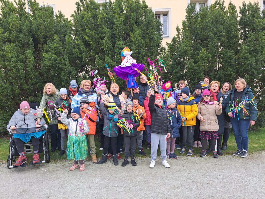 Do zdjęcia pozuje grupa 30 osób -uczniów oraz nauczycieli. Na środku zdjęcia widoczna jest kukła Marzanny. Wszyscy są uśmiechnięci i trzymają w dłoniach gaiki - maiki.