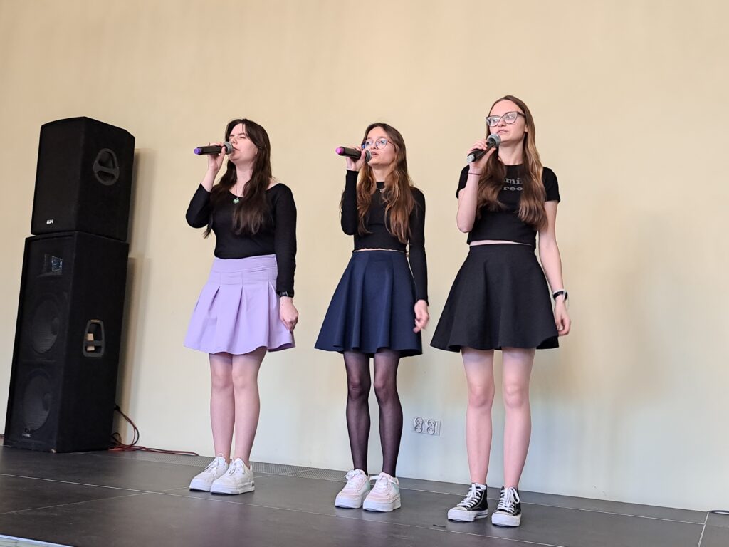 Na scenie stoją trzy dziewczyny. W prawych dłoniach trzymają mikrofon. Ubrane są w czarne bluzki oraz jednokolorowe spódniczki.