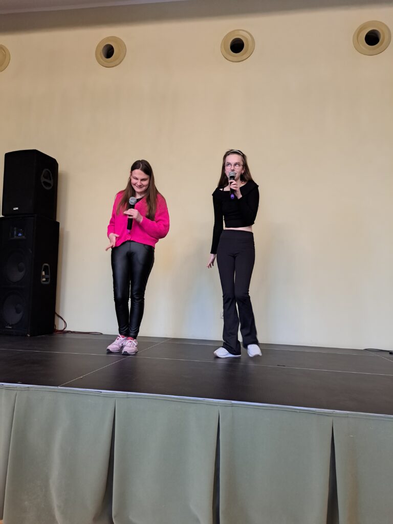 Na scenie stoją dwie dziewczyny ubrane w jednokolorowe bluzki i czarne spodnie.