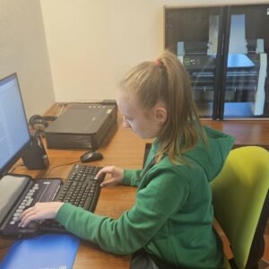 Dziewczyna siedzi przed komputerem i czyta. Na monitorze komputera wyświetlony jest tekst zapisany w wordzie. Dziewczyna trzyma lewą dłoń na monitorze brajlowskim, a prawą dłoń na strzałkach klawiatury. Po prawej stronie dziewczyny znajduje się drukarka brajlowska.