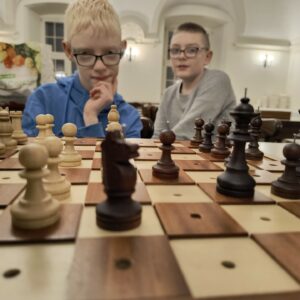 Zdjęcie zrobione w dużej stołówce. Na pierwszym planie znajduje się szachownica. Przy stole siedzi dwóch chłopaków.