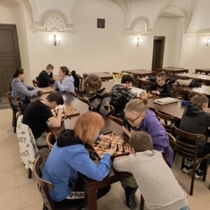 Widok na dużą stołówkę. Przy stołach siedzi dwanaścioro zawodników. Dziewczęta i chłopacy rozgrywają partię szachów.